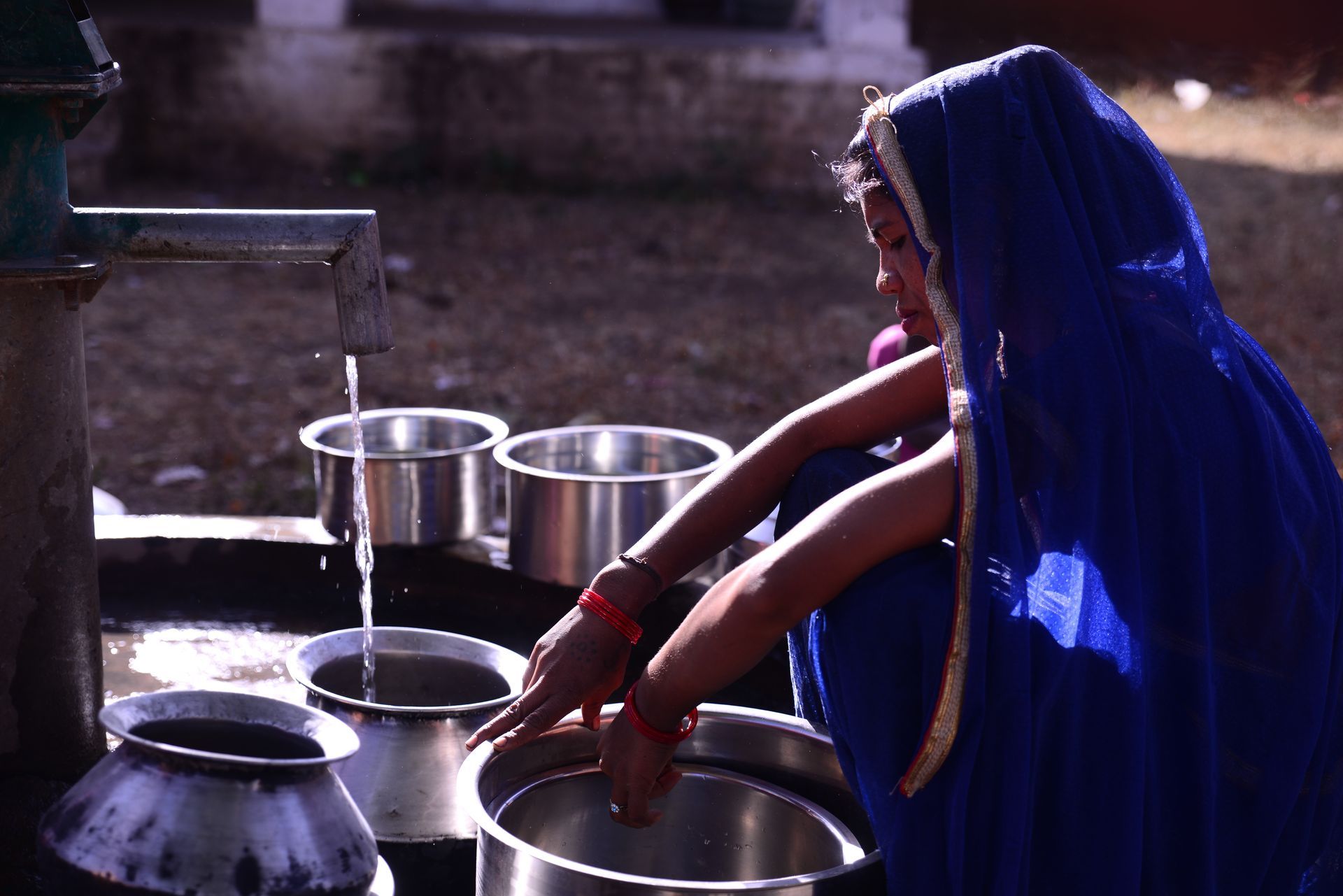 Girl in blue sari at a water pump, filling metal pots in a courtyard.