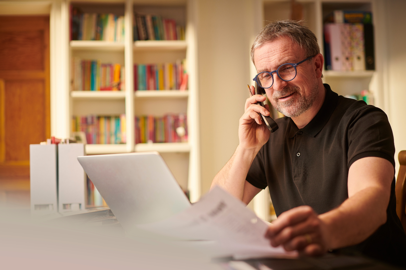 A man is sitting at a desk talking on a cell phone while looking at papers.
