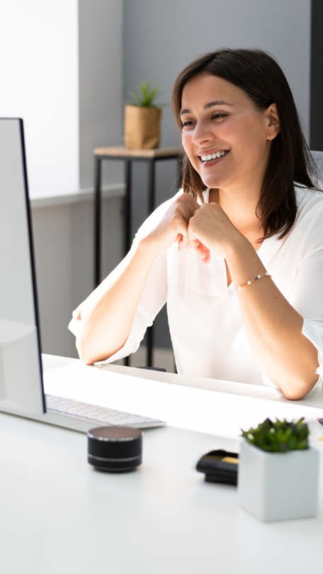 Woman smiling during a video call at her desk, hands clasped, near a computer. Bright office setting.