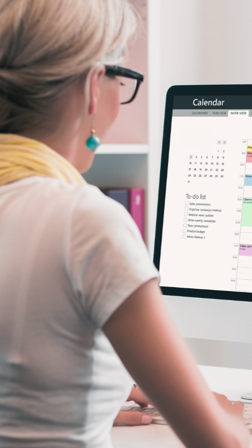 Woman with glasses looking at a computer calendar; white shirt, yellow scarf, and turquoise earrings.