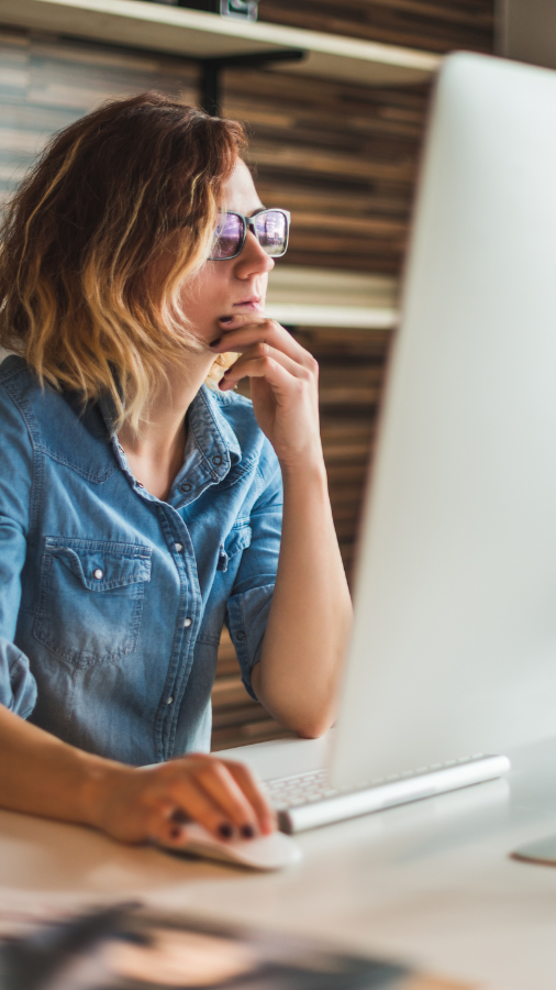 Woman in glasses at a computer, pondering with hand on chin. Wearing denim shirt in an office setting.
