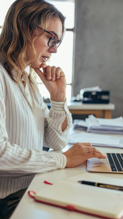 Woman in glasses working on a laptop at a desk, hand on chin, office setting.
