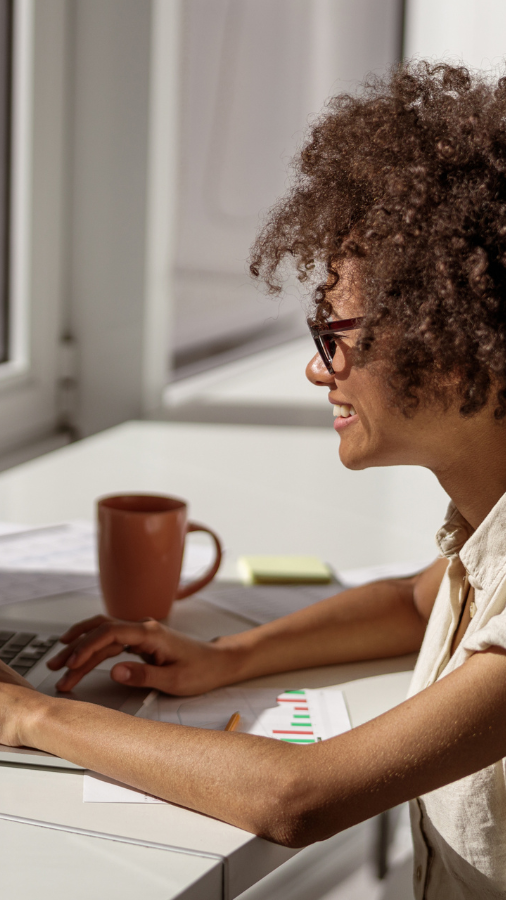 Woman with curly hair smiles while typing on a laptop at a desk, with coffee cup and papers.