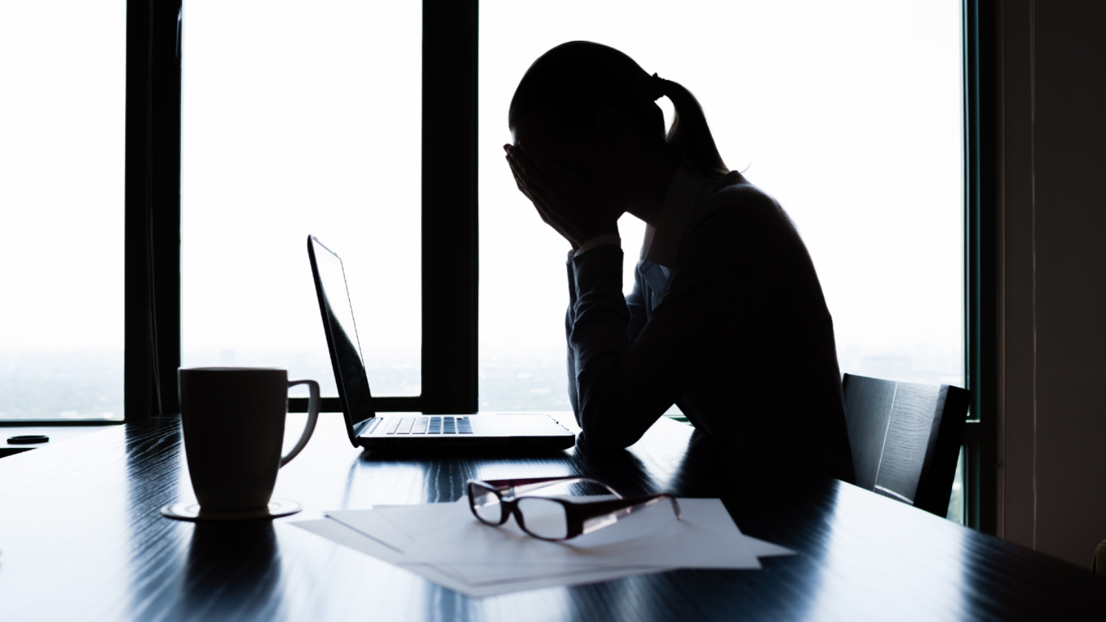 Silhouette of person with head in hands at desk, laptop, papers, and coffee cup nearby.