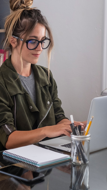 Woman in glasses, working on laptop at a desk with notebook and pencils.