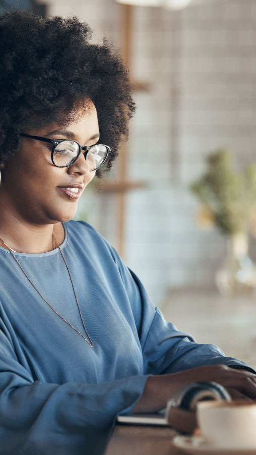 Woman with glasses, curly hair, and blue shirt working on a laptop in a cafe.
