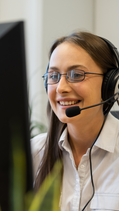 Woman with headset, smiling, wearing glasses, in front of a computer.