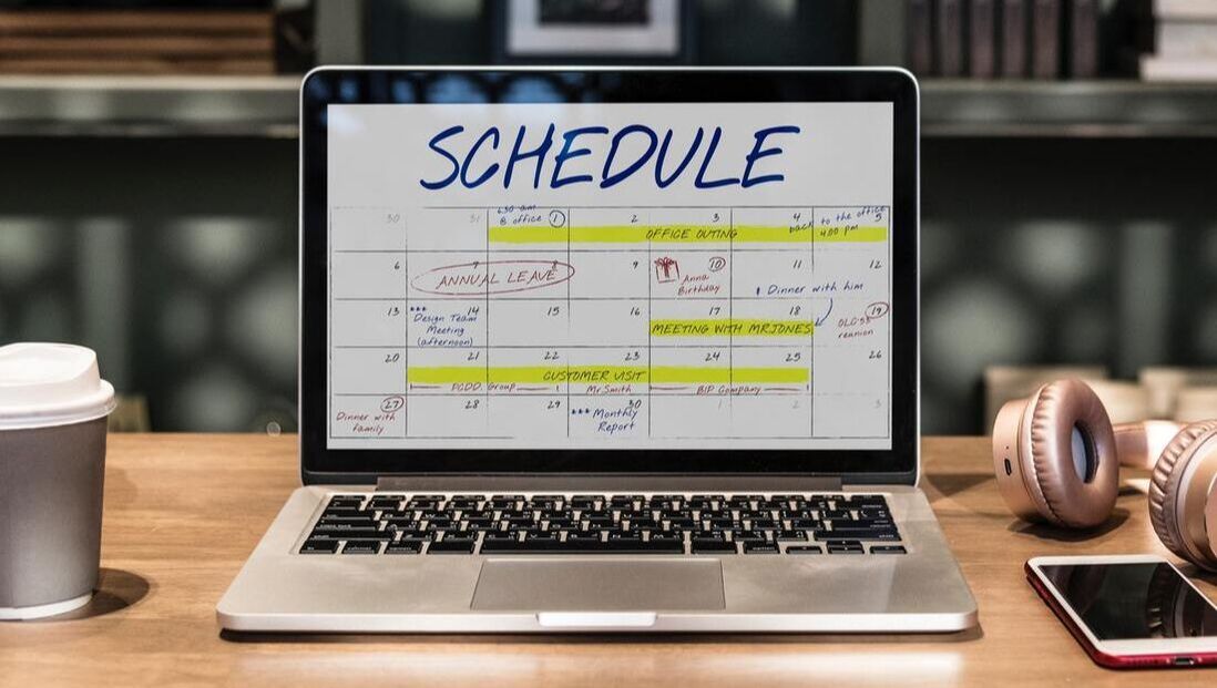 A laptop computer is sitting on a wooden desk with a schedule on the screen.