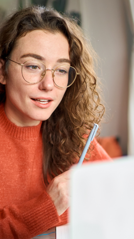 Woman with curly hair wearing glasses and orange sweater looks at laptop, holding pen.