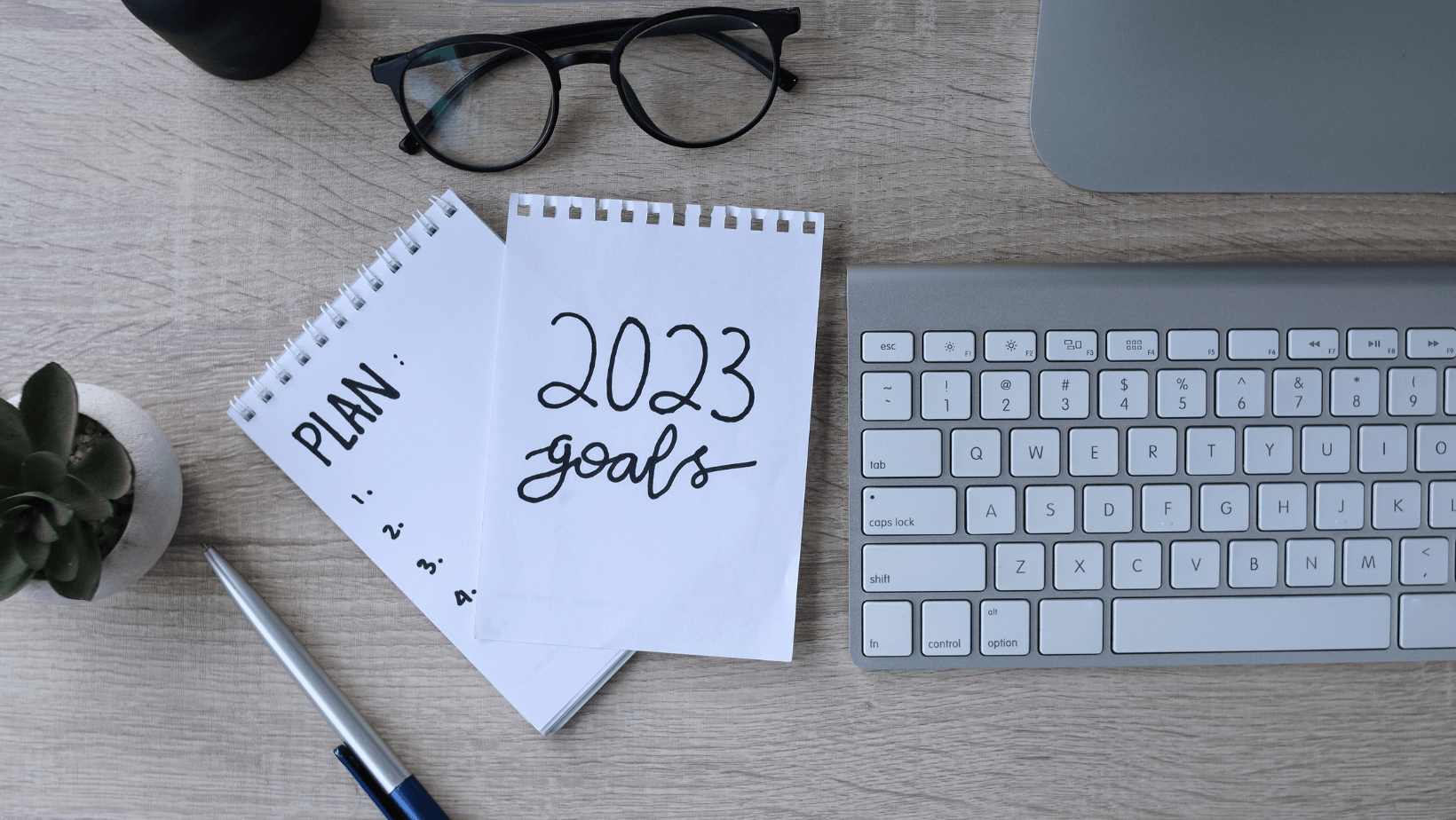 A notepad with the words `` plan 2023 goals '' written on it is on a desk next to a keyboard and glasses.