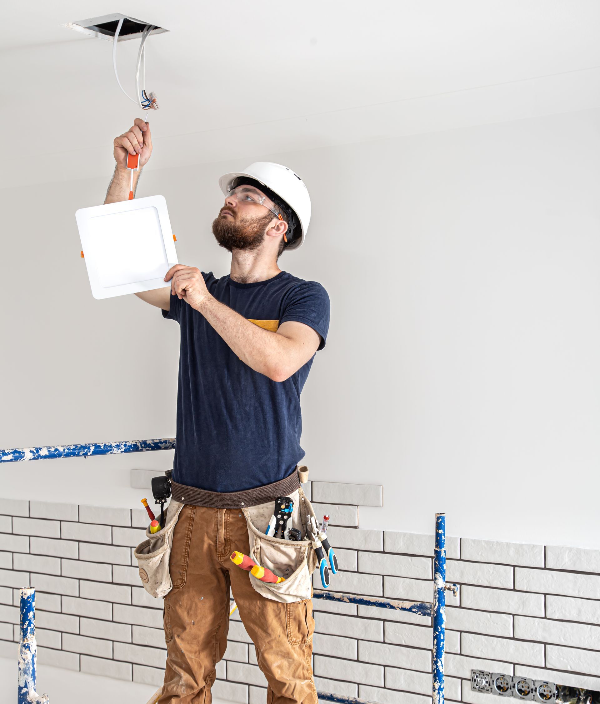electrician-builder-with-beard-worker-white-helmet-work-installation-lamps-height-professional-overalls-with-drill-background-repair-site