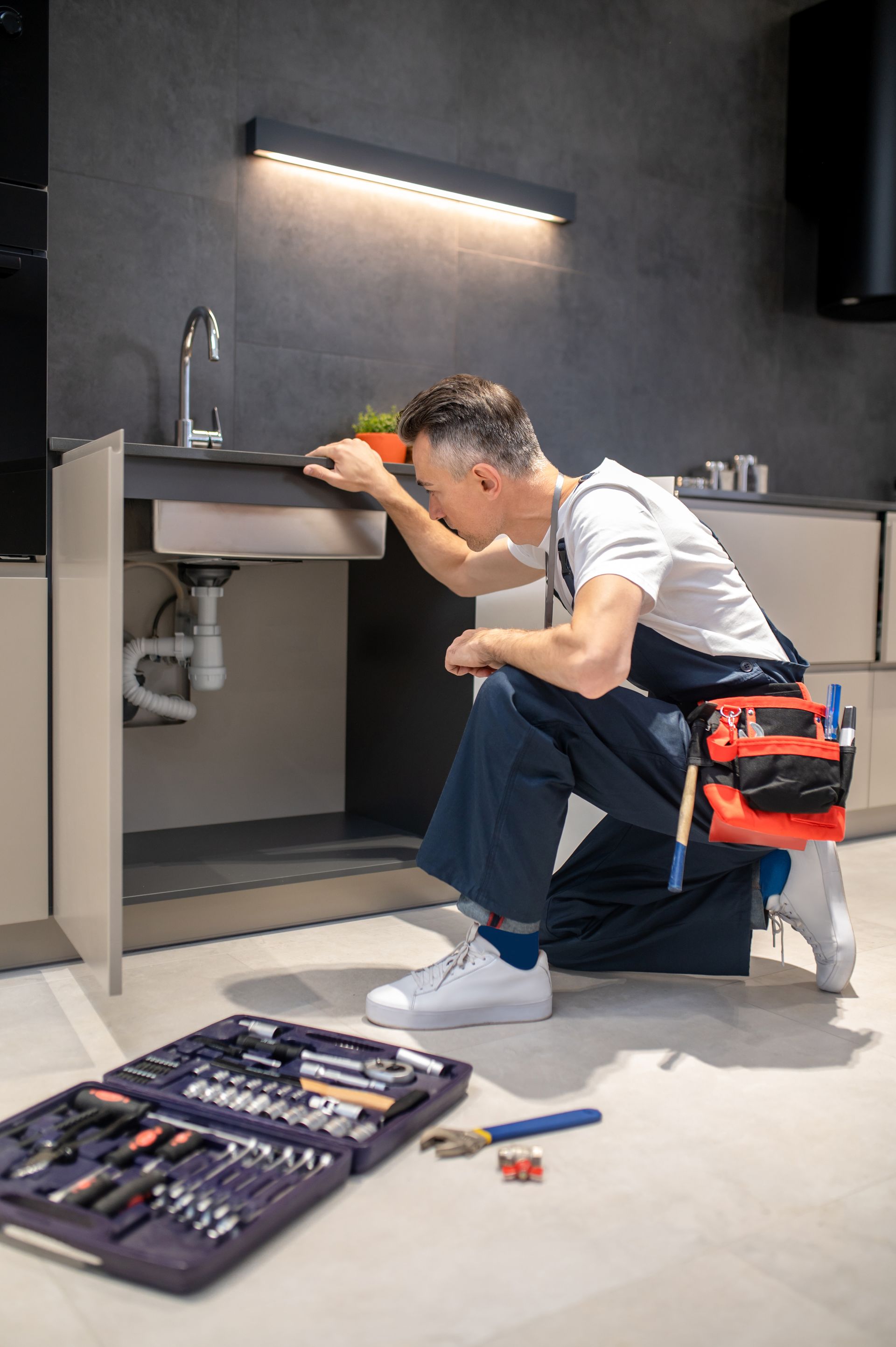 man kneeling down touching sink inspecting pipes