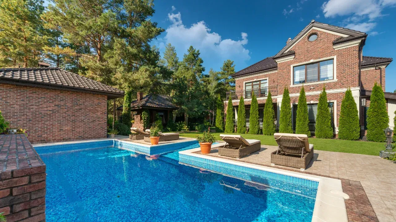 a summer house with tall hedges and a swimming pool with a concrete deck