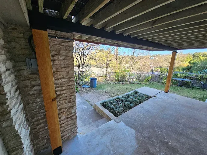 Stone patio with a view, shaded by a wooden pergola with a rectangular planter and surrounding yard.
