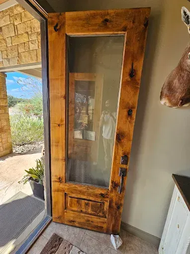 Wooden front door with large window. Person reflected. Moose head on wall, sunny outdoors visible.