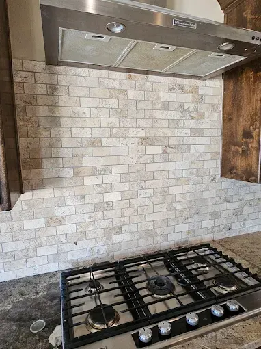 Gas stovetop below a stainless steel range hood and light-colored brick backsplash.