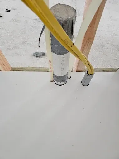 Pipes and wires emerging from a gray cylinder, piercing a white surface, surrounded by wood framing.