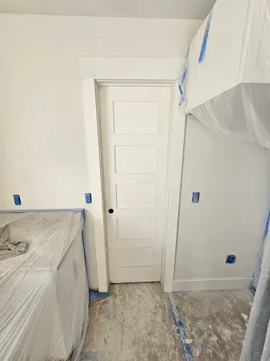 White painted room with a closed door, blue tape and plastic coverings on surfaces.