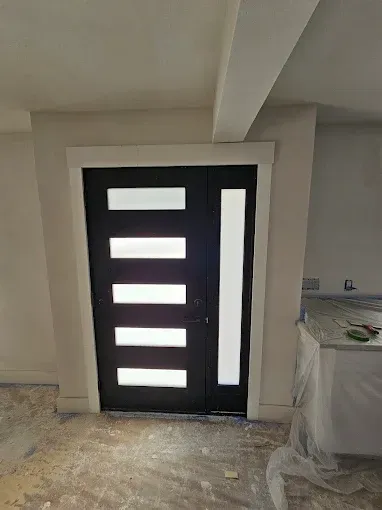Black modern door with frosted glass panels and side window in a room under construction.