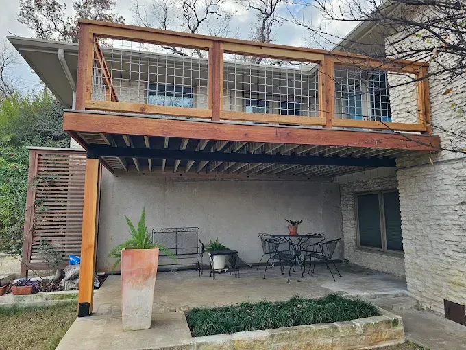 A two-story deck extends from a stone home. The upper level has a wooden railing. The lower level has a patio.