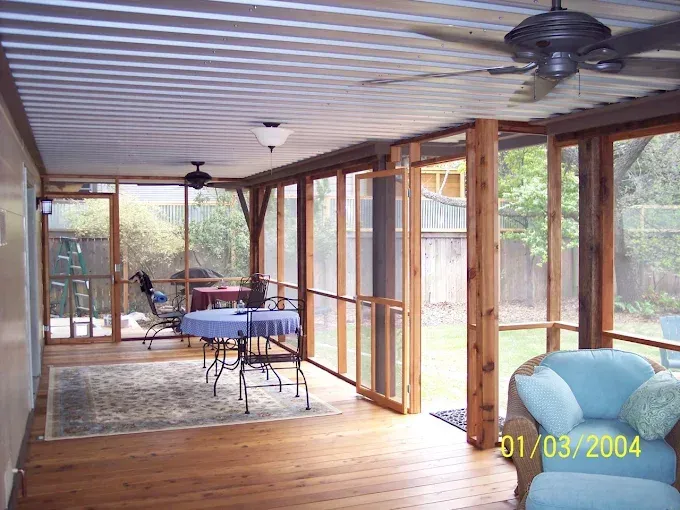 Sunroom with wood frame and screen windows, view to backyard. Blue furniture and a rug on wooden floor.