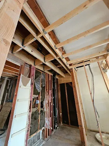 Interior view of a room under construction; exposed wooden beams and studs, wiring, drywall, and an open doorway.