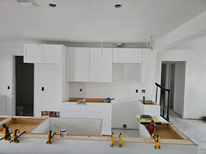 White kitchen cabinets being installed, unfinished, with a wooden countertop base and surrounding walls.