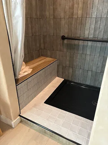 Shower with gray tile, wooden bench, black shower pan, and a grab bar.