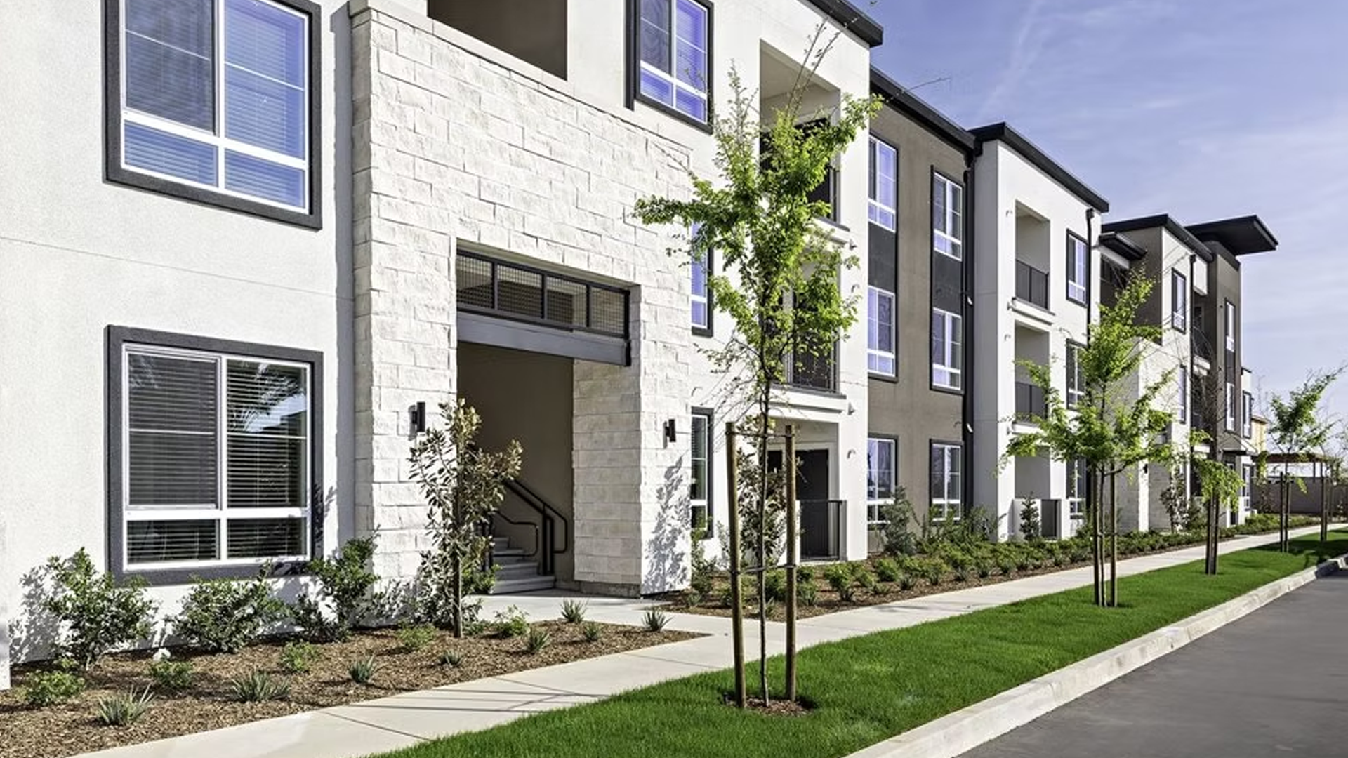 Multi-story apartment building with light-colored exterior and modern design, featuring a walkway and young trees.