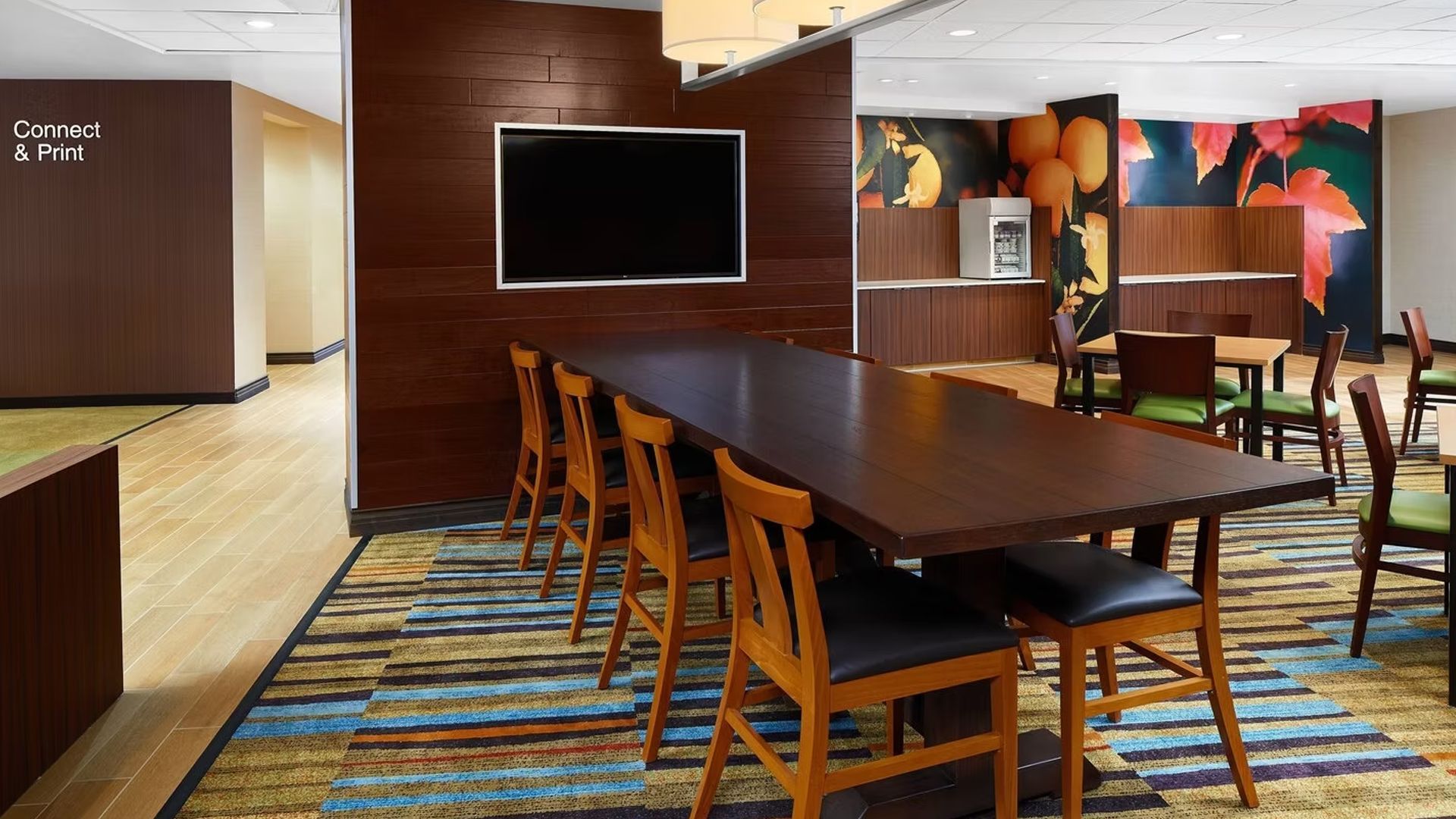 Hotel dining area with long table, TV, and chairs. Brown and gold accents.