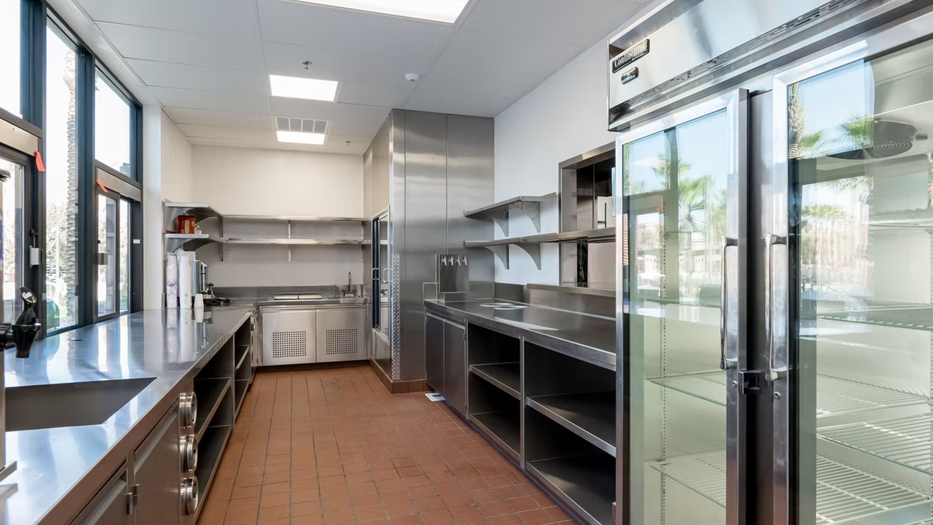 Commercial kitchen with stainless steel appliances and shelving, with windows along the wall.