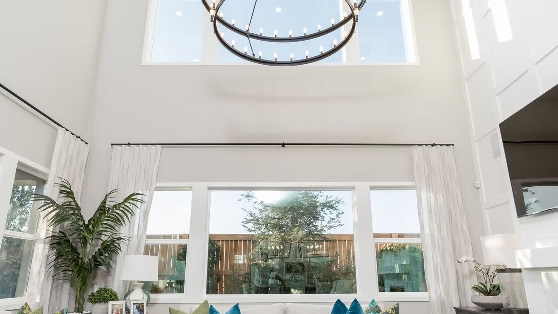 High-ceiling living room with large windows, chandelier, white curtains, and greenery view.