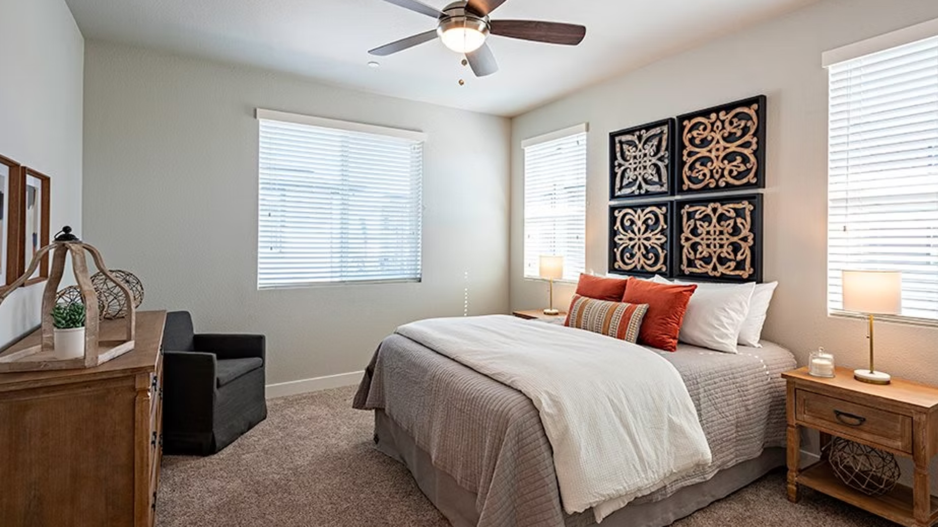 Bedroom with bed, patterned wall decor, two windows with blinds, and wooden furniture.