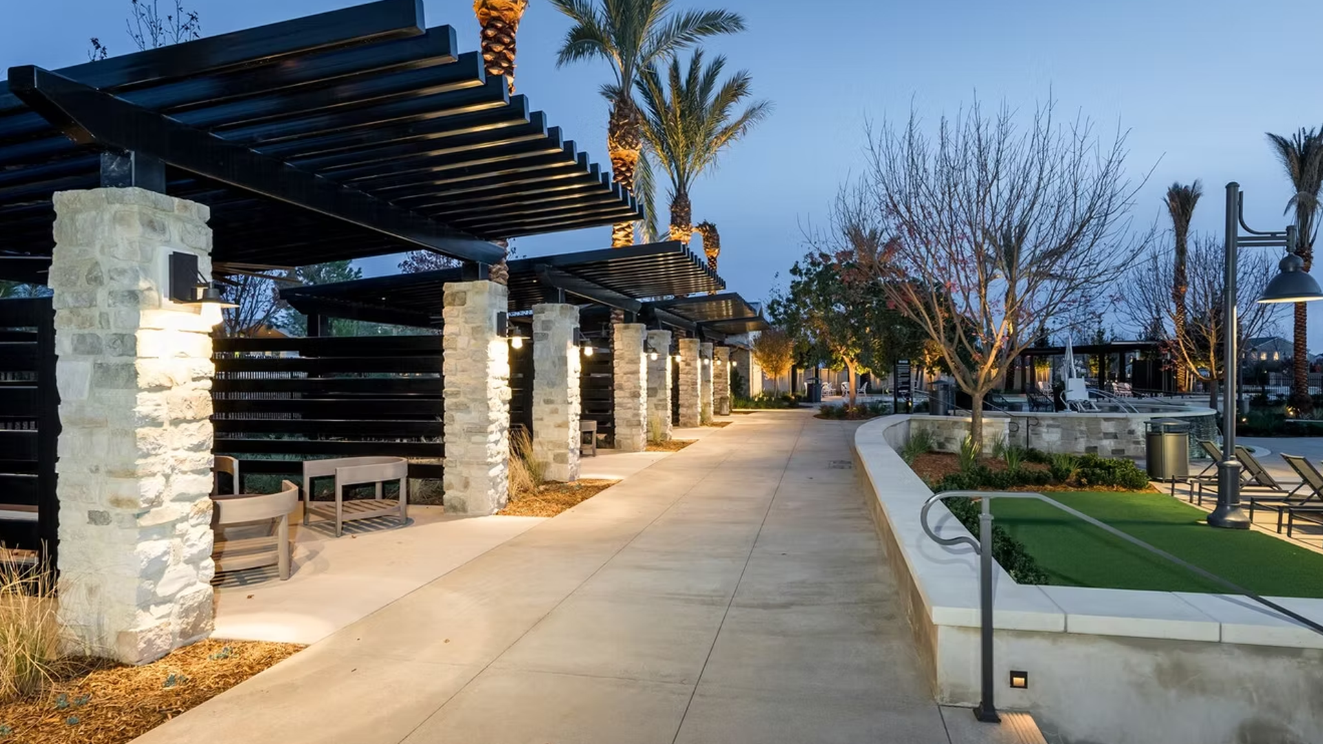 Outdoor walkway lined with stone pillars, a wooden pergola, and landscaping at dusk.