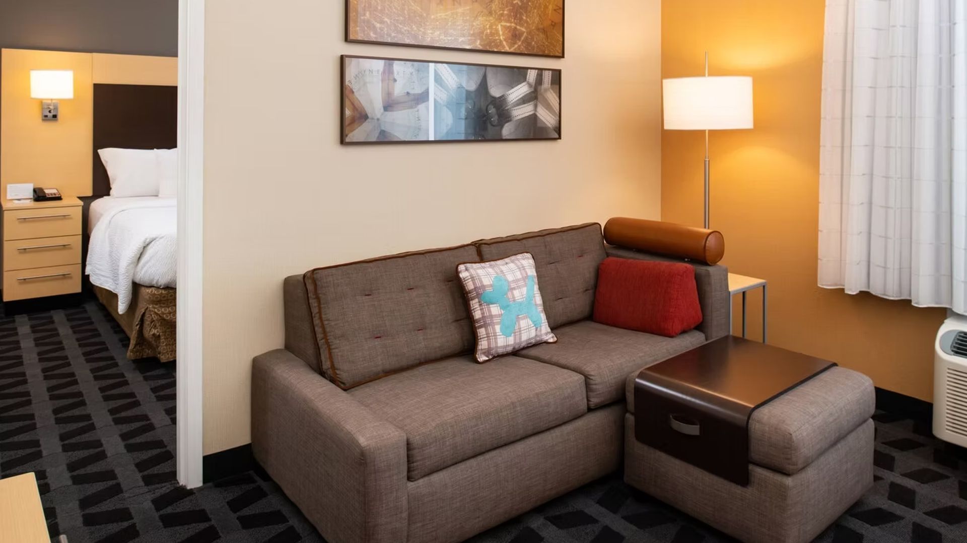 Hotel suite with a sofa, ottoman, and bed visible. Brown, tan, and blue tones.