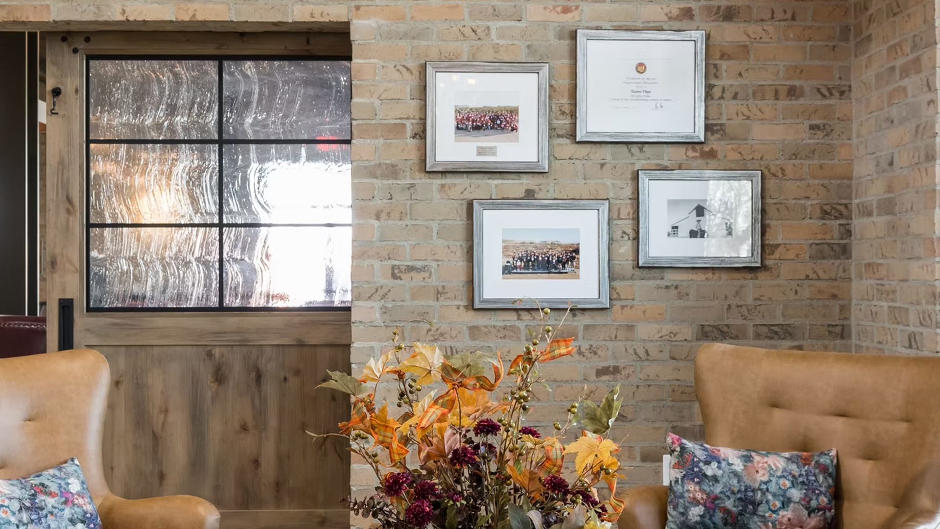 Brick wall with framed art and a window, two leather chairs, and a flower arrangement.