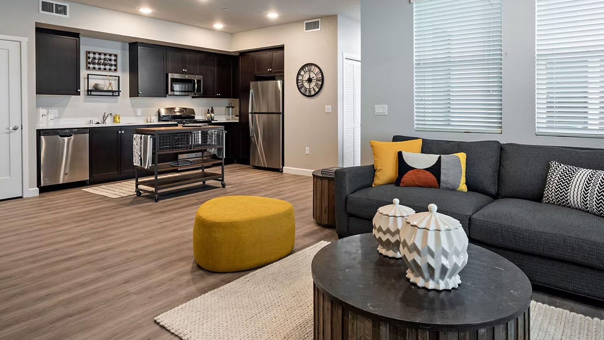 Modern living room and kitchen with dark cabinets, stainless steel appliances, gray sofa, and yellow ottoman.