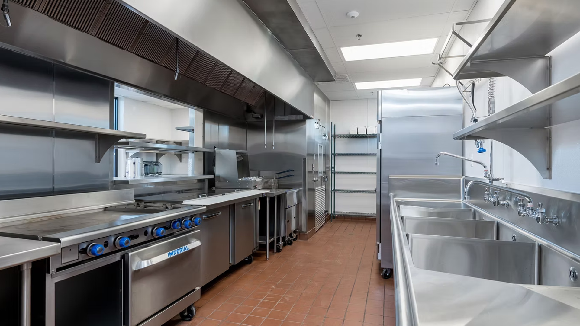 Commercial kitchen with stainless steel appliances: stove, sinks, shelving, and refrigerator.