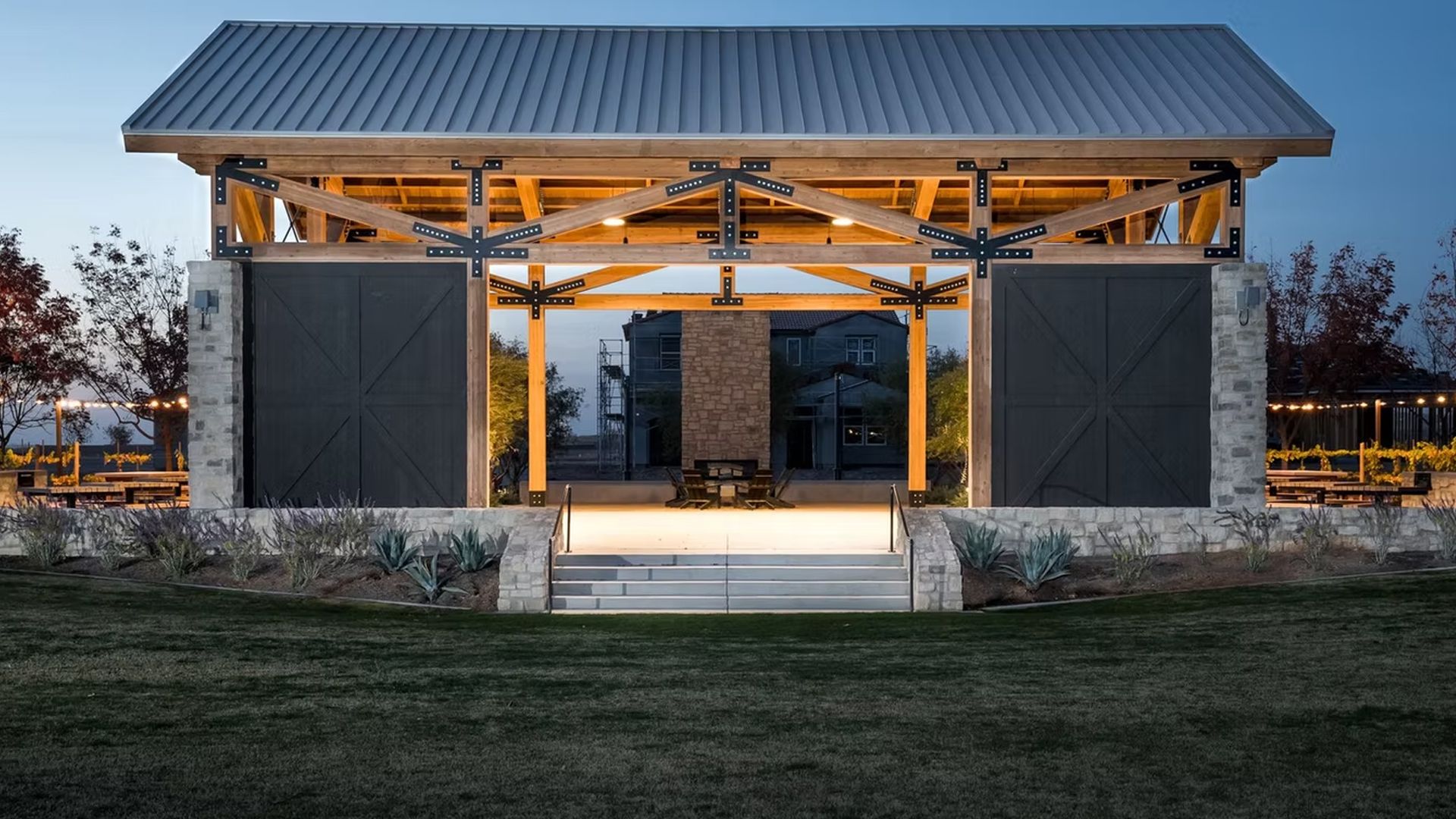 Outdoor stage with stone base, wooden frame, and metal roof, at dusk.