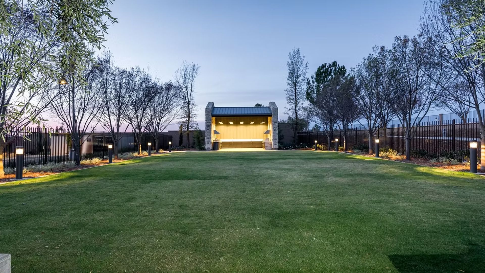 Lush green lawn with trees and a small stone structure at the far end, lit by lampposts.