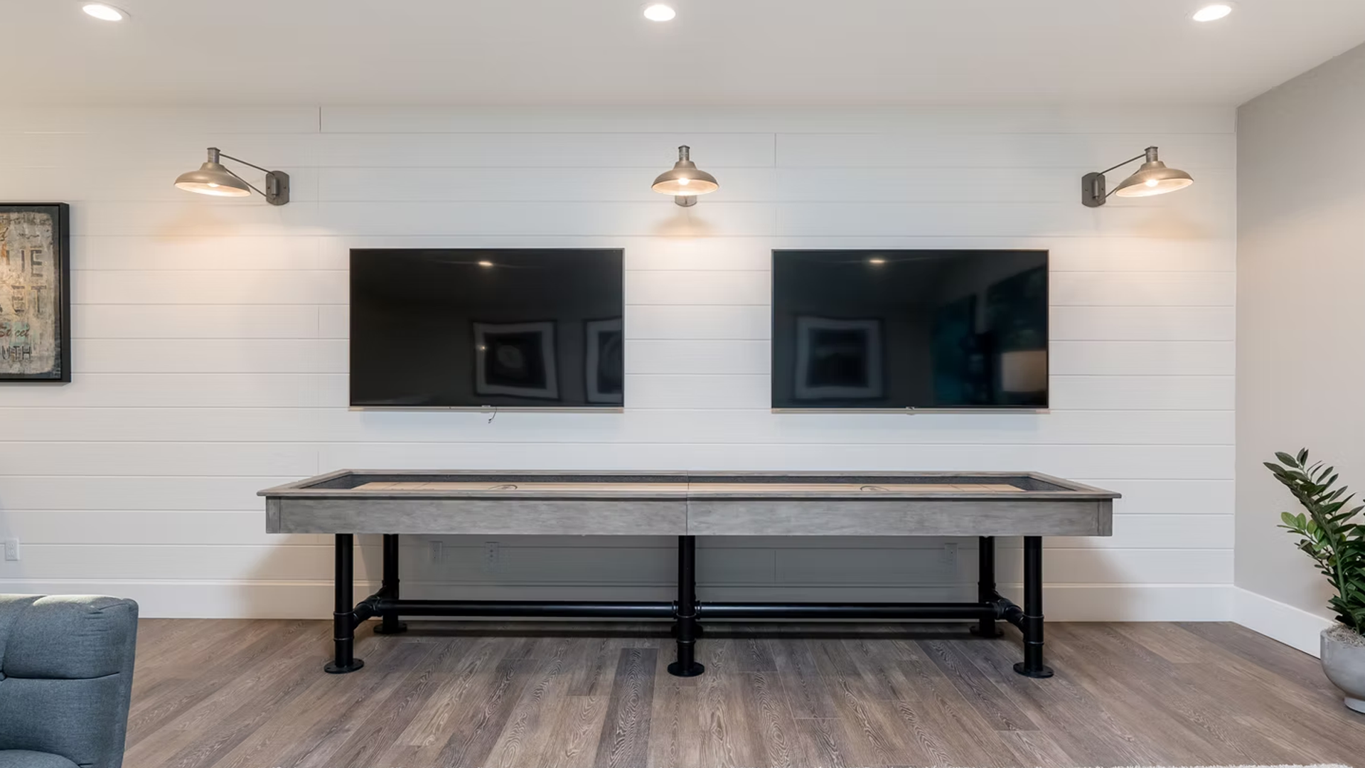 Two TVs mounted on a white shiplap wall above a shuffleboard table. Industrial sconces and a plant on the right.