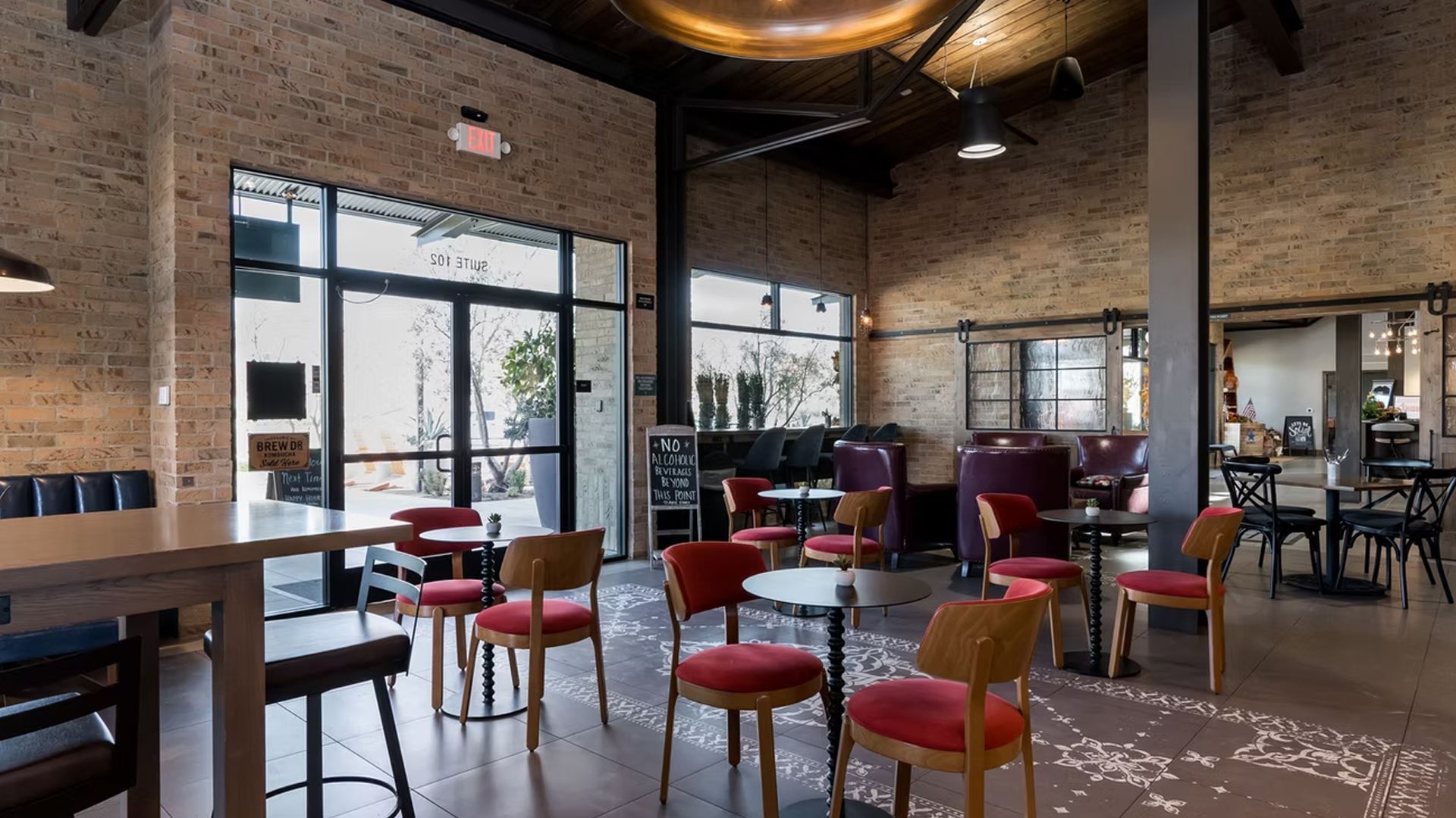 Interior view of a cafe with brick walls, tables, and chairs. Large windows let in light.