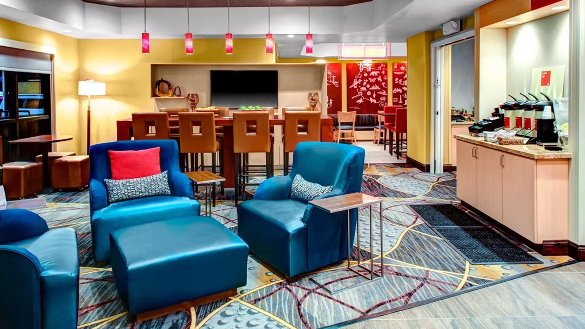 Hotel lobby with seating area and coffee station. Teal armchairs, a large table, and red accents.