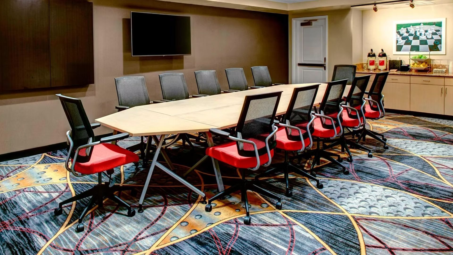 Conference room with long table, black and red chairs, mounted TV, and patterned carpet.
