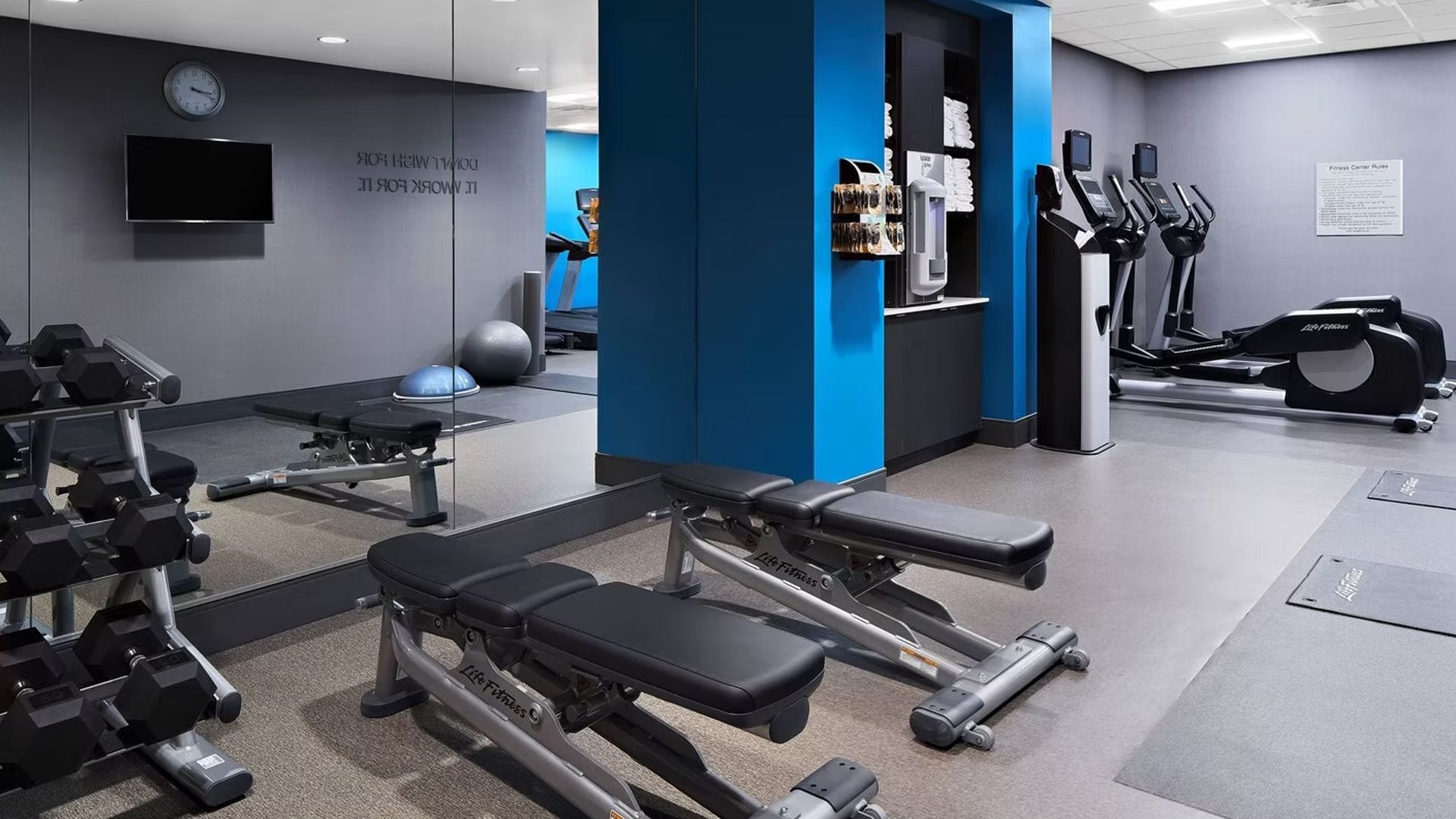 Gym with weights, benches, elliptical, and coffee station; blue and gray walls.