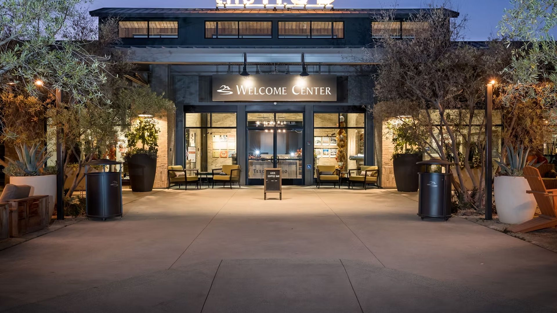 Night view of a Welcome Center entrance, lit with exterior lights. Trees and seating flank the entryway.