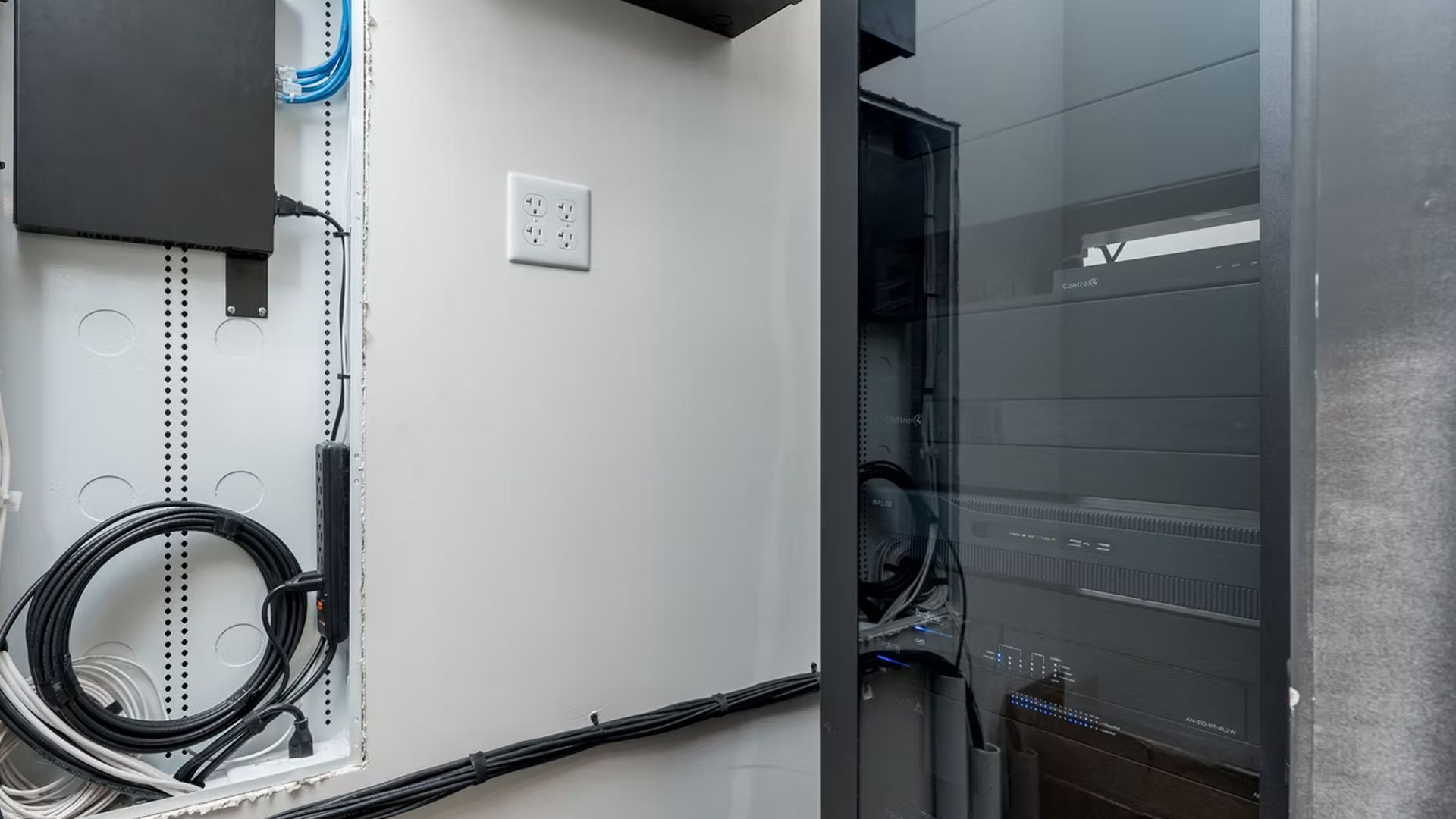 Electrical panel and networking equipment in a white-walled room, black cables, and a glass-doored rack.