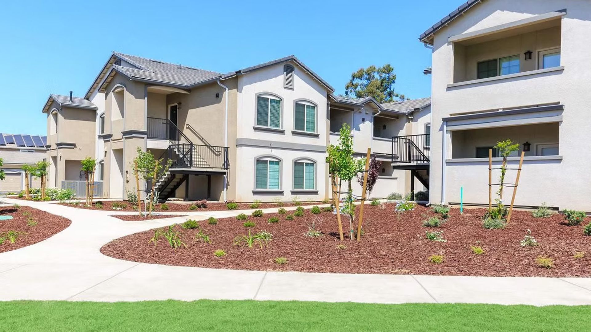 Apartment complex with beige buildings, brown mulch landscaping, and a blue sky.