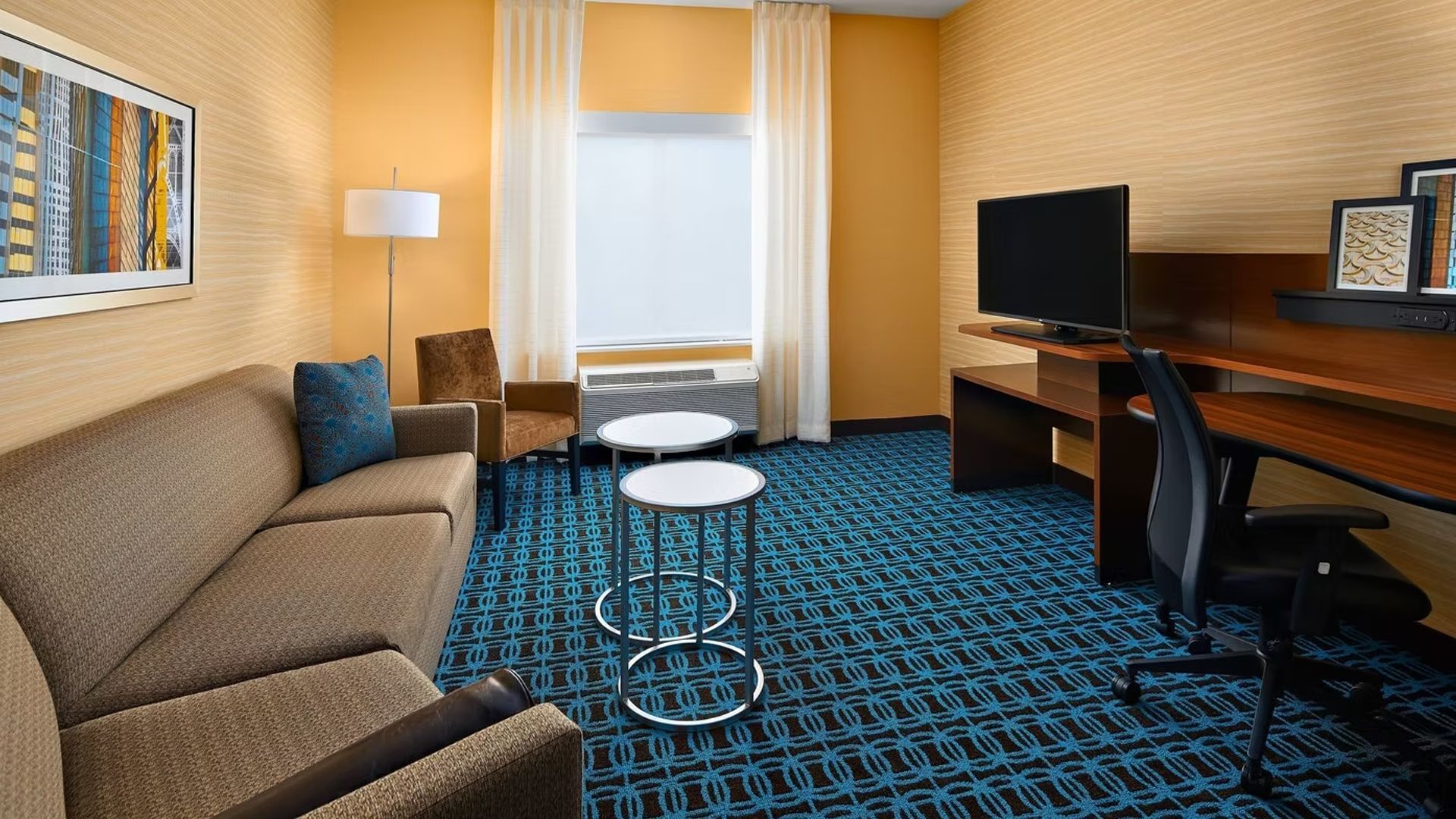 Hotel room with couch, desk, TV, and window; warm colors, blue carpet.