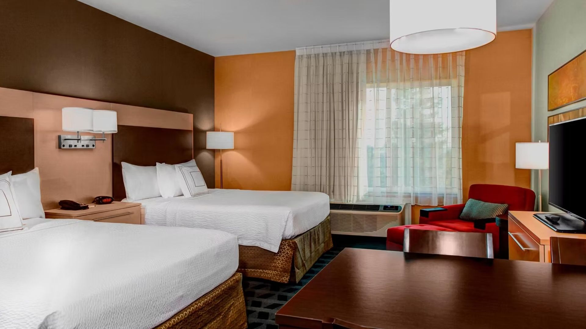 Hotel room with two beds, red armchair, desk, window, and TV.